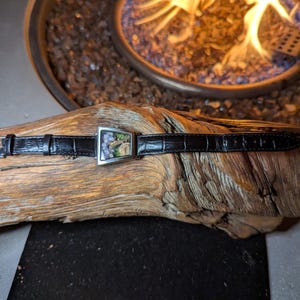 May include: A black leather watch strap with a rectangular silver buckle featuring a landscape design. The watch strap is lying on a piece of driftwood. The background includes a fire pit with flames.