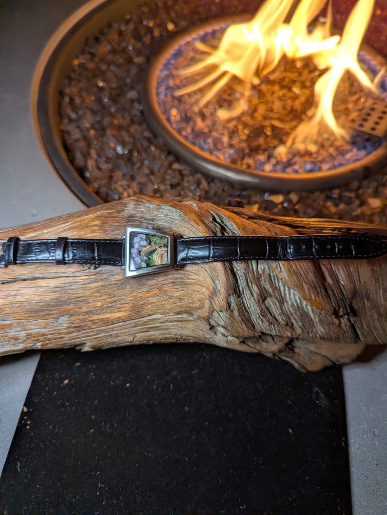 May include: A black leather watch strap with a rectangular silver buckle featuring a landscape design. The watch strap is lying on a piece of driftwood. The background includes a fire pit with flames.