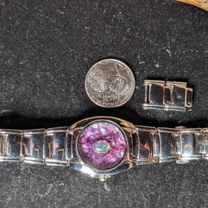 May include: A silver wristwatch with a round face featuring a purple and iridescent stone inlay. The watch is accompanied by a quarter and a watch band link. The watch band is made of silver links.