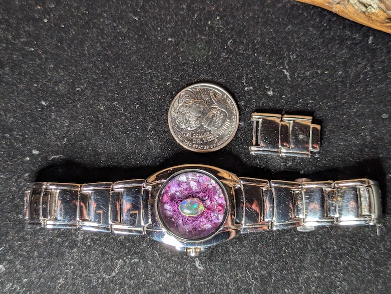 May include: A silver wristwatch with a round face featuring a purple and iridescent stone inlay. The watch is accompanied by a quarter and a watch band link. The watch band is made of silver links.