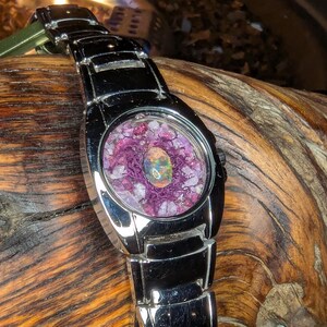 May include: A silver wristwatch with a unique face. The watch face features a central opal surrounded by purple material and small crystals. The watch band is also silver and has a rectangular link design. The watch is resting on a piece of wood.