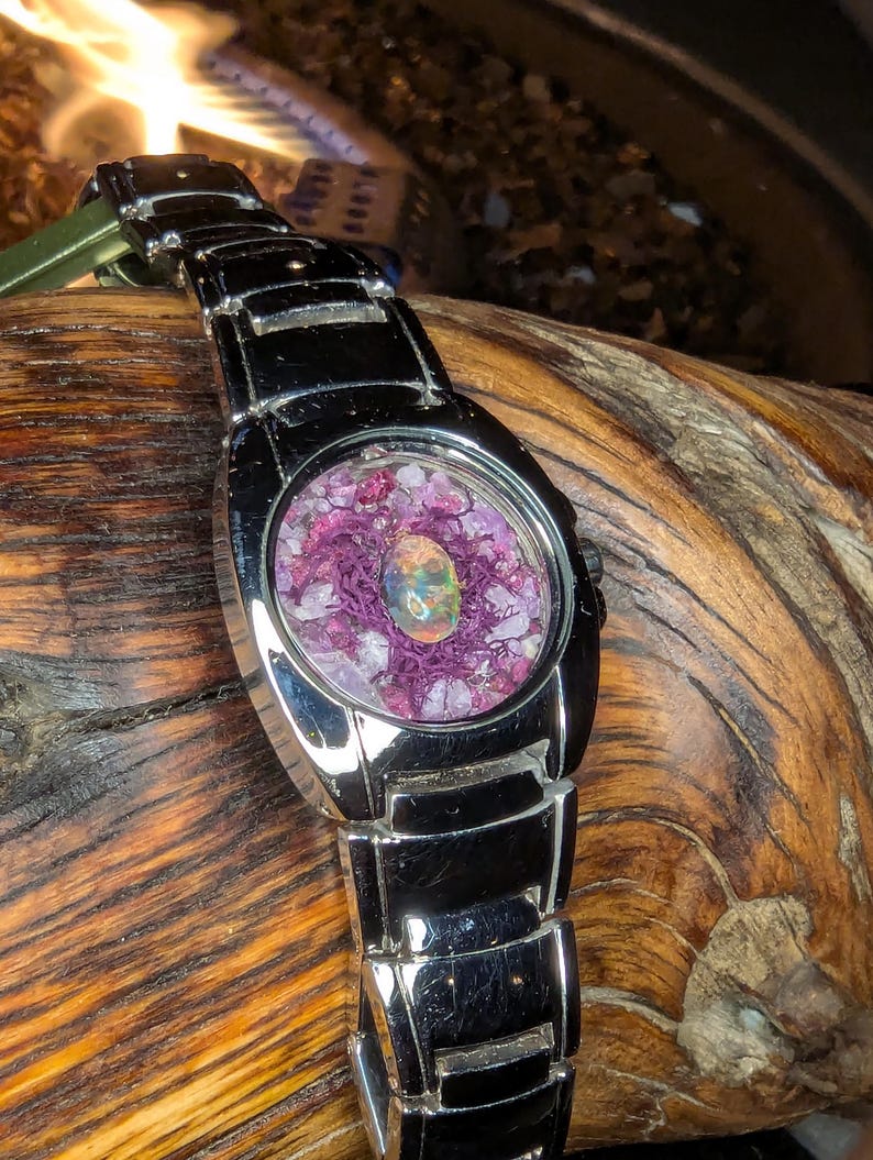 May include: A silver wristwatch with a unique face. The watch face features a central opal surrounded by purple material and small crystals. The watch band is also silver and has a rectangular link design. The watch is resting on a piece of wood.
