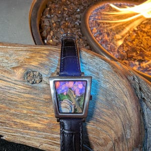 May include: A rectangular wristwatch with a silver case and a black leather strap. The watch face features a colourful floral design. The watch is set against a backdrop of wood and a fire pit.