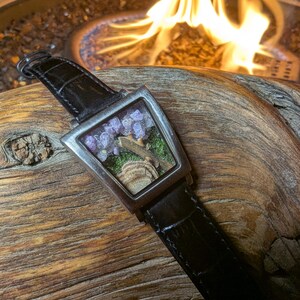 May include: A distinctive wristwatch with a rectangular face, showcasing a miniature landscape with purple flowers and a small rock formation. The watch has a silver bezel and a black leather strap.