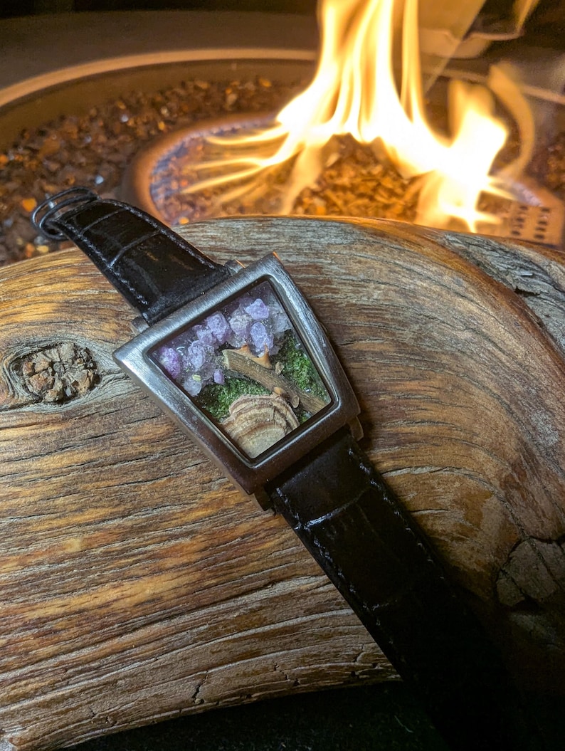 May include: A distinctive wristwatch with a rectangular face, showcasing a miniature landscape with purple flowers and a small rock formation. The watch has a silver bezel and a black leather strap.