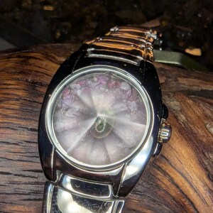 May include: A silver wristwatch with a black bezel and a unique face. The watch face features a circular design with pink and purple accents. The watch band is made of silver links, and the watch is displayed on a wooden surface.