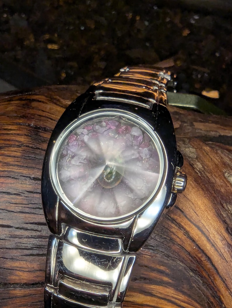May include: A silver wristwatch with a black bezel and a unique face. The watch face features a circular design with pink and purple accents. The watch band is made of silver links, and the watch is displayed on a wooden surface.