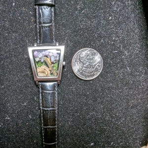 May include: A distinctive wristwatch with a black leather strap and a silver-coloured, triangular watch face. The watch face displays a miniature landscape with purple and green elements. A coin is next to the watch for size comparison.