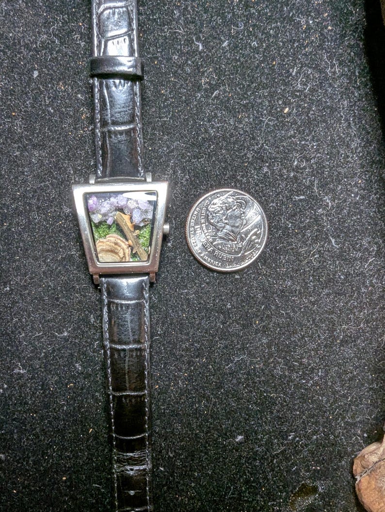 May include: A distinctive wristwatch with a black leather strap and a silver-coloured, triangular watch face. The watch face displays a miniature landscape with purple and green elements. A coin is next to the watch for size comparison.