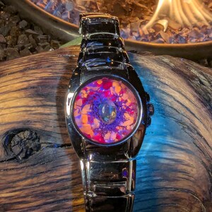 May include: A stainless steel wristwatch with a unique face design. The watch face features a colorful, abstract pattern with shades of purple, orange, and pink. The watch is set against a dark wooden surface.