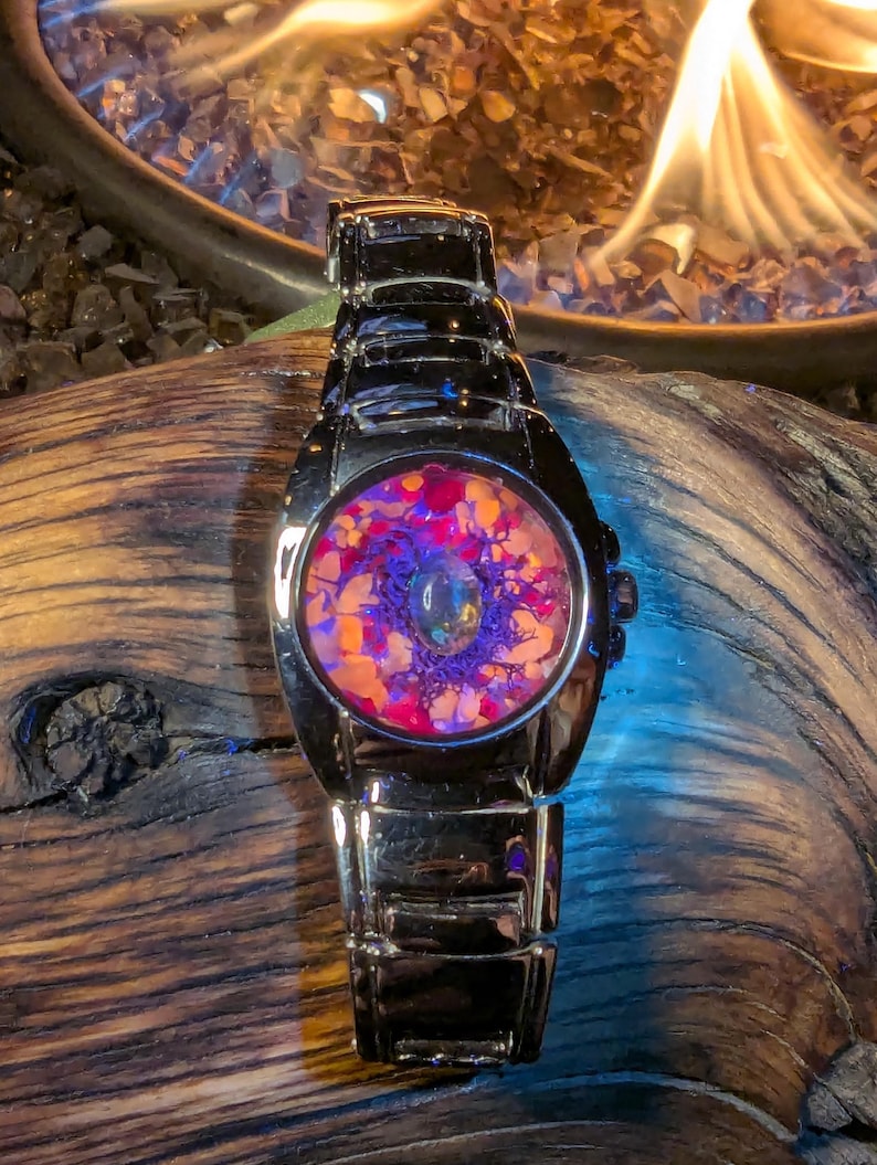 May include: A stainless steel wristwatch with a unique face design. The watch face features a colorful, abstract pattern with shades of purple, orange, and pink. The watch is set against a dark wooden surface.