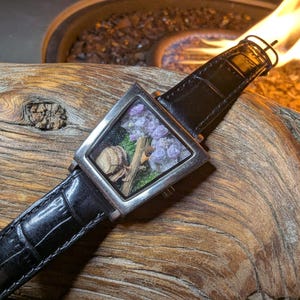 May include: A square wristwatch with a black leather strap. The watch face is a clear square containing small purple flowers, green moss, and a small piece of wood. The watch is set against a textured wooden surface.