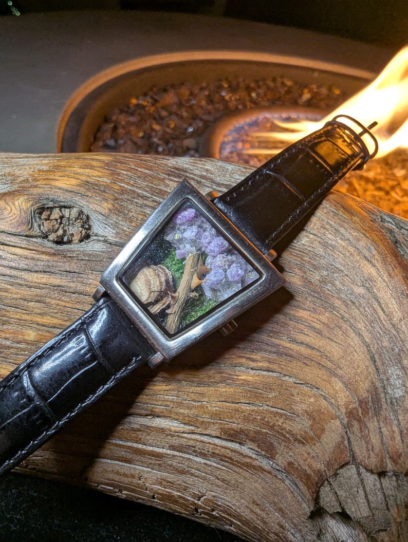 May include: A square wristwatch with a black leather strap. The watch face is a clear square containing small purple flowers, green moss, and a small piece of wood. The watch is set against a textured wooden surface.