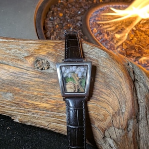 May include: A distinctive wristwatch with a square, silver-toned face, featuring a miniature terrarium scene with rocks and greenery. The watch has a black leather strap with a crocodile pattern. The watch is displayed on driftwood.