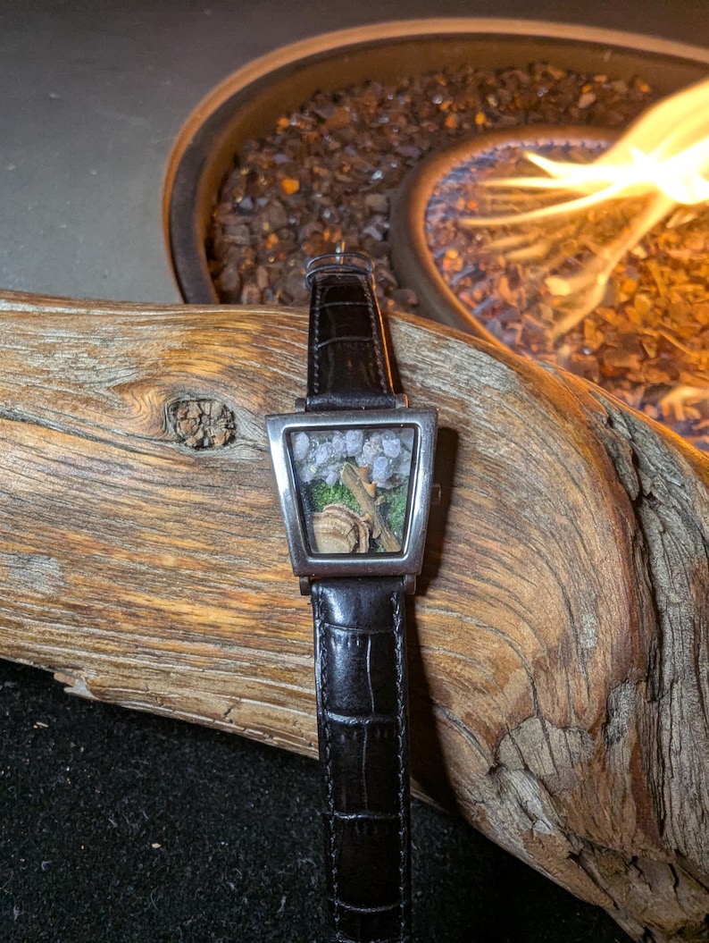 May include: A distinctive wristwatch with a square, silver-toned face, featuring a miniature terrarium scene with rocks and greenery. The watch has a black leather strap with a crocodile pattern. The watch is displayed on driftwood.