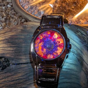 May include: A black wristwatch with a circular face filled with colorful, abstract elements. The face features a mix of orange, red, and purple hues, with a clear, rounded crystal in the center. The watch has a shiny, metallic band.