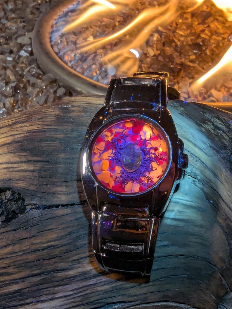May include: A black wristwatch with a circular face filled with colorful, abstract elements. The face features a mix of orange, red, and purple hues, with a clear, rounded crystal in the center. The watch has a shiny, metallic band.