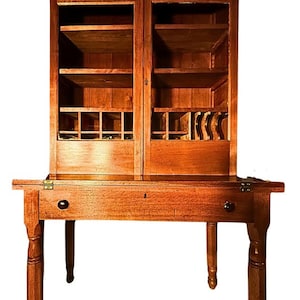 May include: A wooden antique cabinet with a desk. The cabinet has two glass-paned doors and multiple shelves. The desk has a drawer and is supported by four legs. The wood has a warm, reddish-brown tone.