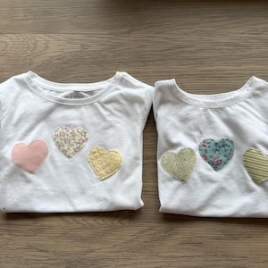 May include: Two white t-shirts with heart appliqués. One shirt has a pink, floral, and yellow heart. The other shirt has a gold glitter, floral, and green striped heart. The shirts are from Old Navy.