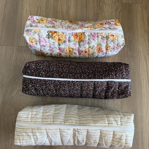 May include: Three quilted, zippered bolster pillows. The top pillow has a white background with yellow and pink roses. The middle pillow is dark brown with small floral patterns. The bottom pillow is cream with gold stripes.