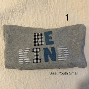 May include: A gray pillow with the words "BE KIND" appliqued on the front. The letters are made of various fabrics, including denim, gingham, and striped patterns. The size is Youth Small.