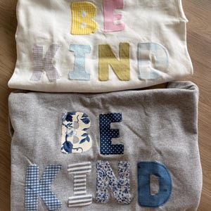 Könnte beinhalten: Zwei Sweatshirts, eines in Weiß und eines in Grau, jeweils mit dem Schriftzug BE KIND aus verschiedenen gemusterten Stoffen appliziert. Das weiße Sweatshirt hat pastellfarbene Buchstaben, das graue zeigt Buchstaben in Blautönen.