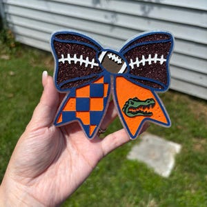May include: A decorative bow with a football design. The bow features a brown glitter top section with white stitching, a football in the center, and orange and blue checkered and solid sections. A green alligator graphic is on the orange section.