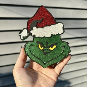 May include: A green Grinch-themed decoration with a red glitter Santa hat and yellow eyes. The Grinch's face is outlined in black, and the hat has white trim. The decoration is held in a hand.
