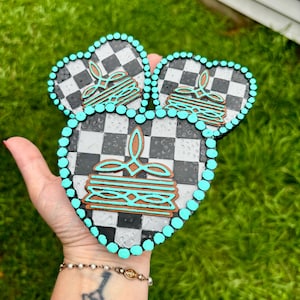 May include: A set of three heart-shaped coasters with a black and white checkered pattern. Each coaster features a brown and turquoise pancake design. The edges are outlined with turquoise beads. The coasters are held in a hand.