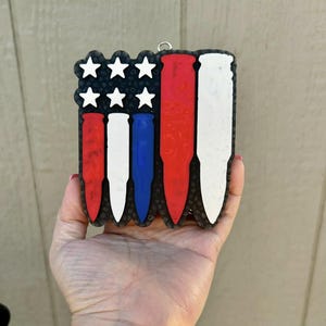 May include: A patriotic-themed ornament shaped like the American flag, featuring red, white, and blue bullet shapes. The ornament has white stars in the upper left corner and a black border. It is held in a hand.
