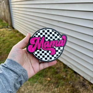 May include: A round, black and white checkered design with the word "Mama" in pink glitter. The design is on a black background.