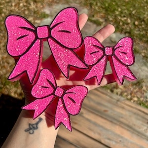 May include: Three pink glitter bows with black outlines are displayed. The bows vary in size, with the largest at the top and the smallest at the bottom. The bows are a vibrant pink color and have a glittery texture.