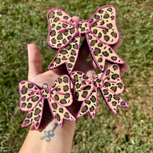 May include: A collection of five leopard print bows with pink trim. The bows are in various sizes and have a small metal ring at the top for hanging. The bows are set against a background of green grass.