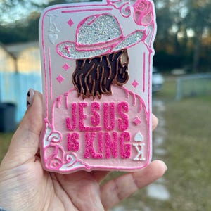 May include: A pink and white playing card-shaped decoration with the words "JESUS IS KING" in glittery pink letters. The design features a figure wearing a hat with glitter and brown hair. A pink rose and the letter "K" are also visible.