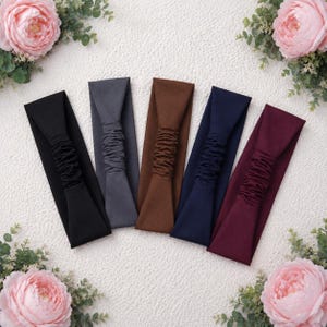 Può includere: Cinque fasce per capelli in tessuto disposte in fila, dotate di una sezione elastica arricciata al centro. I colori da sinistra a destra sono nero, grigio antracite, marrone, blu navy e bordeaux scuro. Ogni fascia ha una finitura opaca e liscia.