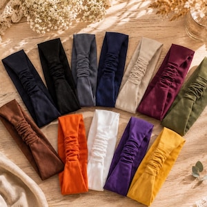 Può includere: Una collezione di dodici fasce per capelli in tessuto in vari colori a tinta unita, tra cui nero, blu navy, grigio, beige, bordeaux, verde oliva, marrone, arancione, bianco, viola e giallo, ciascuna con una sezione elastica arricciata.