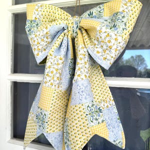 May include: A large, decorative bow made of patchwork fabric, featuring yellow floral, blue floral, and yellow gingham patterns. The bow is suspended by a brown string, and is displayed against a white window.