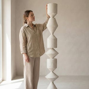 May include: A tall, abstract sculpture with a stacked design of geometric shapes. The sculpture is made of a light-colored material and stands on a dark, circular base. The sculpture's height is approximately 6 feet.
