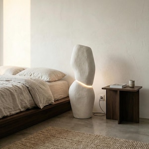 Puede incluir: Un dormitorio minimalista con una l&aacute;mpara de pie blanca abstracta. La l&aacute;mpara tiene una secci&oacute;n iluminada y est&aacute; junto a una mesita de noche de madera oscura con un libro y una taza. Una cama con ropa de cama de tonos neutros y una alfombra tejida completan la escena.