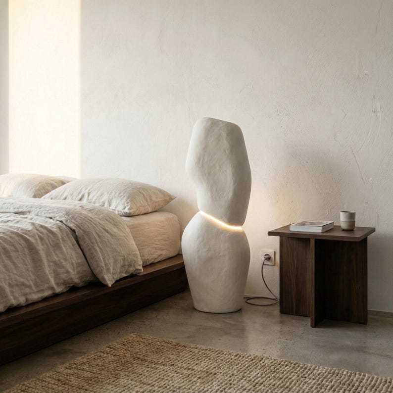 Puede incluir: Un dormitorio minimalista con una l&aacute;mpara de pie blanca abstracta. La l&aacute;mpara tiene una secci&oacute;n iluminada y est&aacute; junto a una mesita de noche de madera oscura con un libro y una taza. Una cama con ropa de cama de tonos neutros y una alfombra tejida completan la escena.