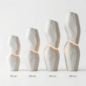 Puede incluir: Cuatro esculturas abstractas de cer&aacute;mica blanca de diferentes alturas, cada una con una banda horizontal de luz brillante. Las esculturas est&aacute;n etiquetadas con sus alturas: 90 cm, 120 cm, 150 cm y 180 cm. Las formas tienen una superficie rugosa y texturizada.