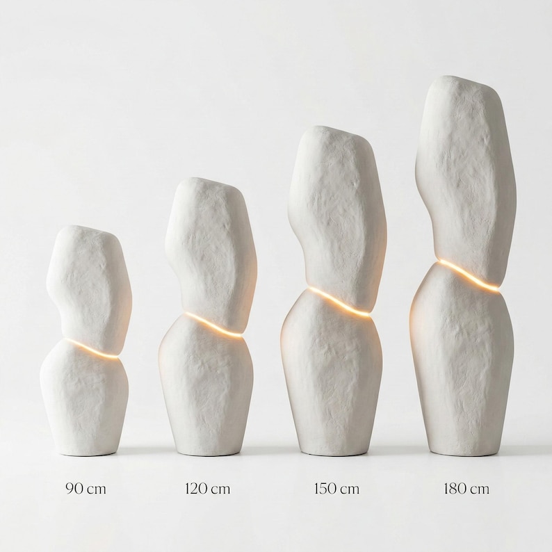 Puede incluir: Cuatro esculturas abstractas de cer&aacute;mica blanca de diferentes alturas, cada una con una banda horizontal de luz brillante. Las esculturas est&aacute;n etiquetadas con sus alturas: 90 cm, 120 cm, 150 cm y 180 cm. Las formas tienen una superficie rugosa y texturizada.