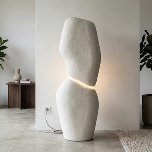 Puede incluir: Una l&aacute;mpara de pie escultural alta, de color blanquecino, con una banda horizontal brillante. La l&aacute;mpara tiene una superficie texturizada y est&aacute; colocada en un sal&oacute;n moderno. La habitaci&oacute;n incluye plantas en macetas y una paleta de colores neutros.