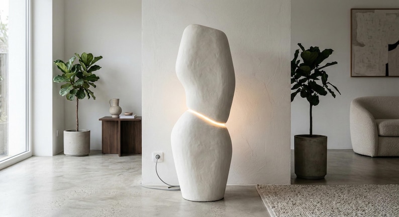 Puede incluir: Una l&aacute;mpara de pie escultural alta, de color blanquecino, con una banda horizontal brillante. La l&aacute;mpara tiene una superficie texturizada y est&aacute; colocada en un sal&oacute;n moderno. La habitaci&oacute;n incluye plantas en macetas y una paleta de colores neutros.