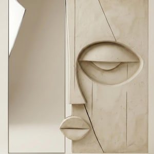 May include: Abstract wall art depicting a sculpted face with a single eye, nose, and lips, in a neutral beige tone. A rectangular mirror is positioned to the left. The design is minimalist and modern.