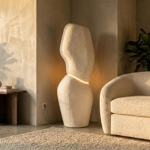 Puede incluir: Un sal&oacute;n moderno con una gran l&aacute;mpara de pie abstracta. La l&aacute;mpara de color crema emite una luz c&aacute;lida, colocada junto a un sof&aacute; curvo de color crema. Una planta en maceta y una mesa auxiliar con decoraci&oacute;n completan la escena.