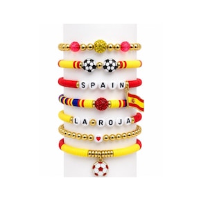 May include: A collection of colorful beaded bracelets. The bracelets feature gold, red, yellow, and blue beads, with soccer ball charms, the word "SPAIN", and a Spanish flag charm. Some bracelets have heart-shaped beads.