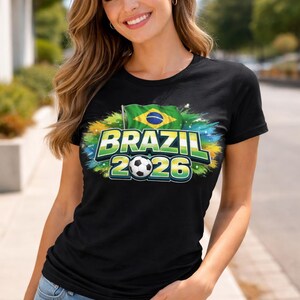 May include: Black t-shirt featuring a Brazil 2026 design. The graphic includes the Brazilian flag, a football, and the text "BRAZIL 2026" in green and blue. The shirt is worn by a person.