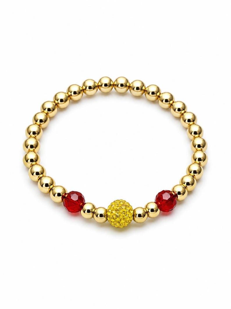 May include: Gold beaded bracelet with a central yellow crystal ball and two red crystal beads. The bracelet is made of small, round, gold-colored beads. The design is simple and elegant.