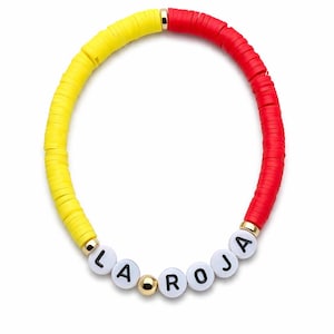 May include: A colorful bracelet featuring yellow and red flat beads. The bracelet includes white letter beads that spell out "LA ROJA" and gold-colored accents. The bracelet is a vibrant accessory.